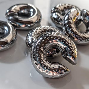 May include: Close-up of several shiny, silver-coloured, donut-shaped ear weights. The weights have a hammered texture and a curved design. The objects are arranged on a white surface, reflecting light.