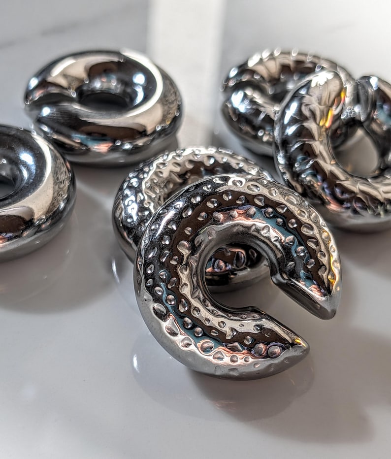 May include: Close-up of several shiny, silver-coloured, donut-shaped ear weights. The weights have a hammered texture and a curved design. The objects are arranged on a white surface, reflecting light.