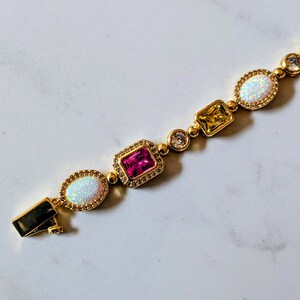 May include: Gold-toned bracelet featuring a series of colorful gemstones. The bracelet includes oval-shaped opals, a rectangular pink stone, a square yellow stone, and round clear stones. The clasp is visible on the left side.