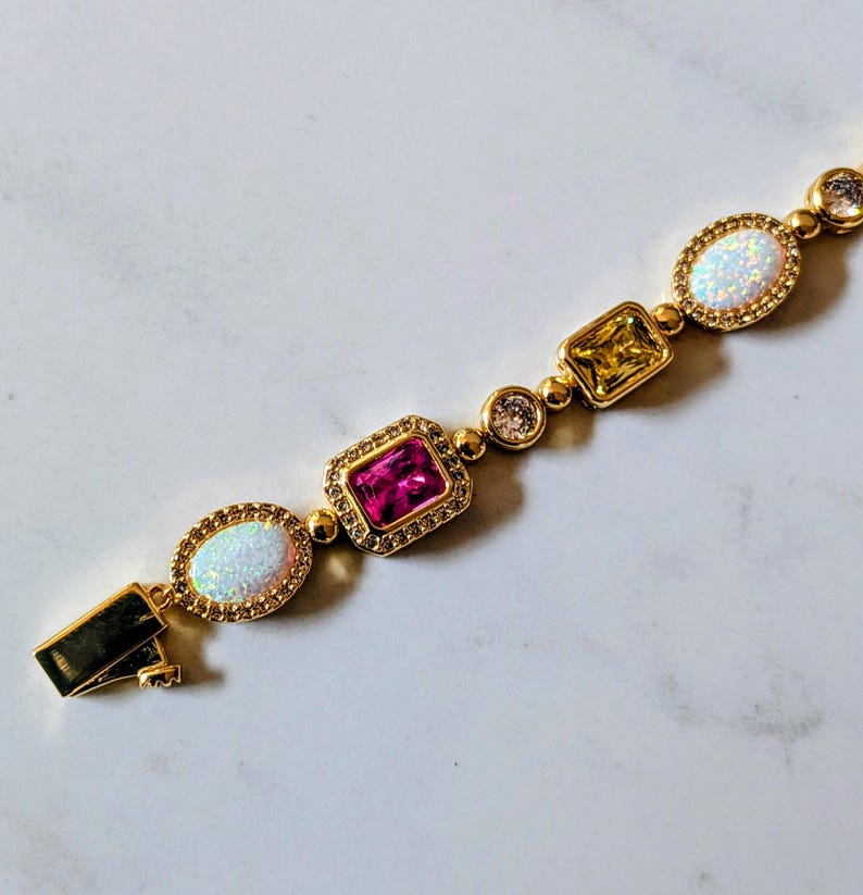 May include: Gold-toned bracelet featuring a series of colorful gemstones. The bracelet includes oval-shaped opals, a rectangular pink stone, a square yellow stone, and round clear stones. The clasp is visible on the left side.