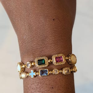May include: Two gold-toned bracelets with colorful gemstones. One bracelet features a green, pink, and opal-like stone. The other bracelet has a blue cross and blue rectangular stone. Both bracelets have a delicate design.