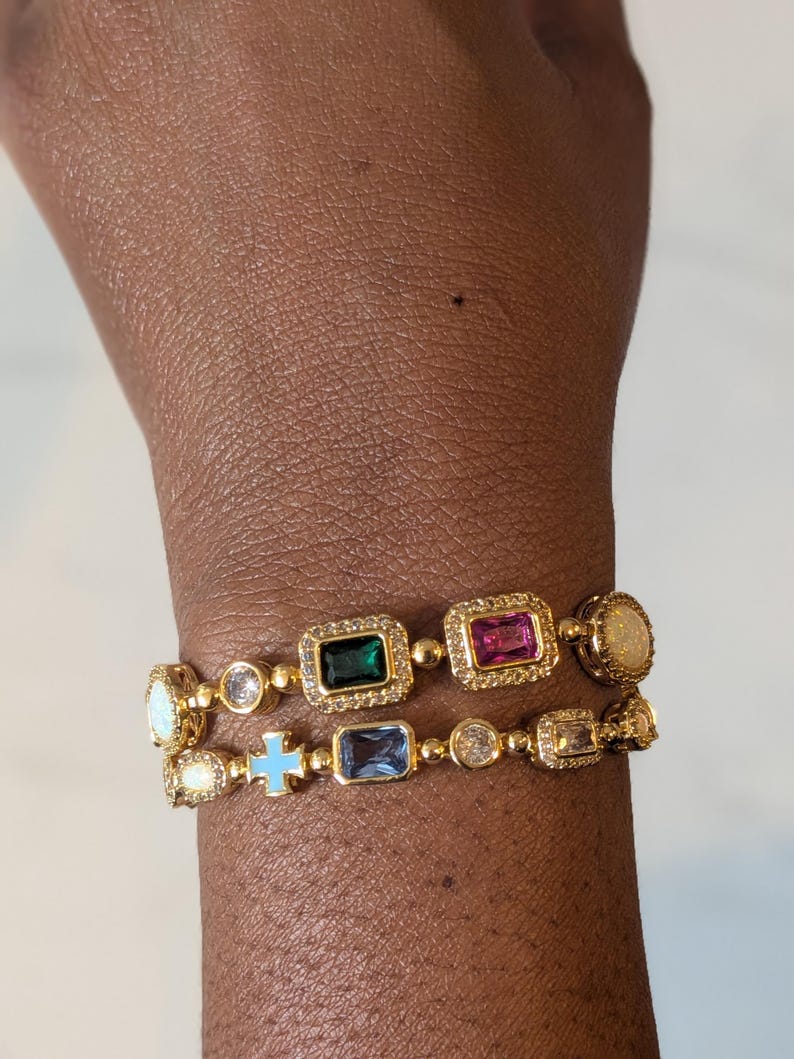May include: Two gold-toned bracelets with colorful gemstones. One bracelet features a green, pink, and opal-like stone. The other bracelet has a blue cross and blue rectangular stone. Both bracelets have a delicate design.