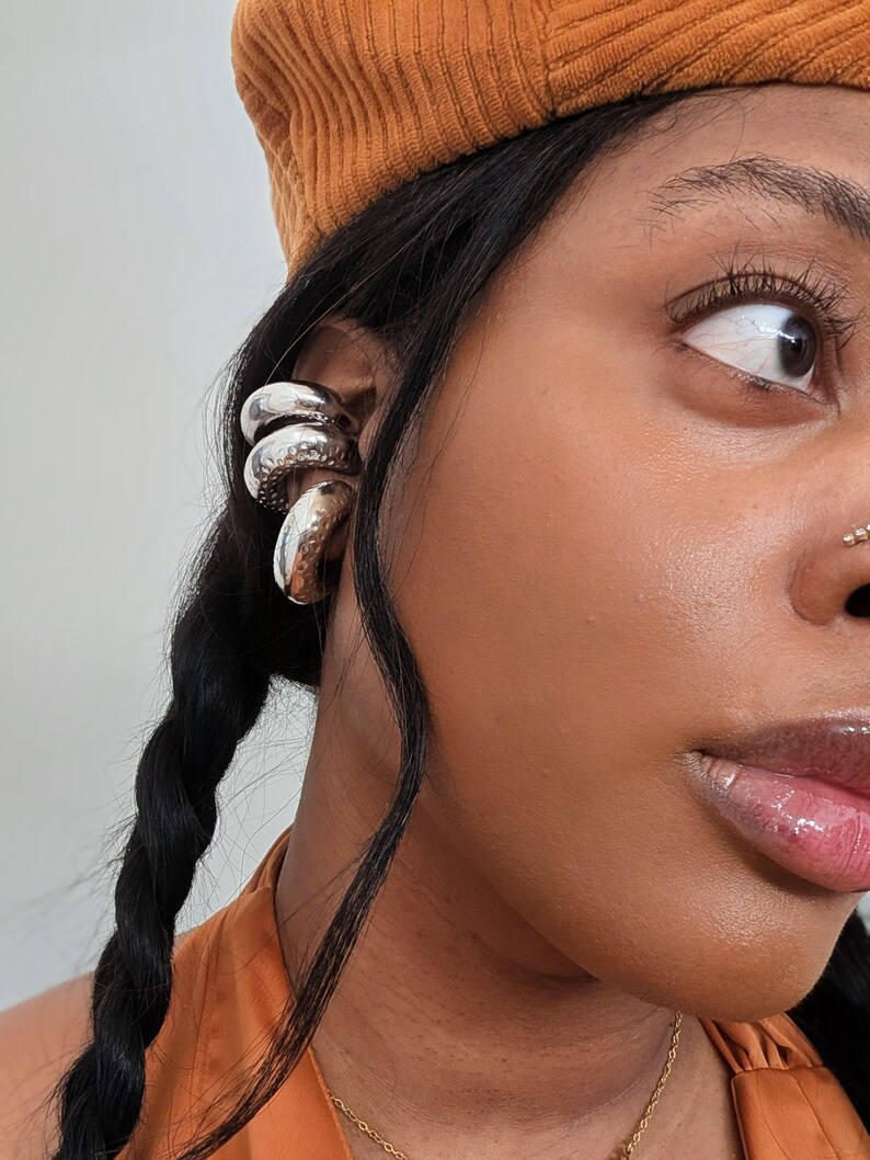 May include: Silver ear cuffs with a hammered texture are worn on the ear. The person is wearing an orange beret and an orange top. A gold necklace is visible. The person has dark hair in a braid.
