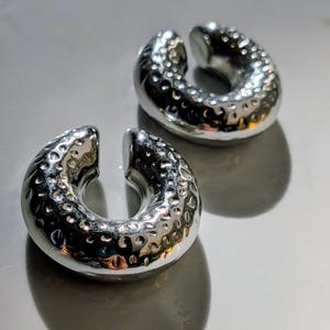 May include: Two silver ear weights with a hammered texture. The weights are circular with a split opening, designed for stretched earlobes. They are displayed on a reflective surface. Each weight is approximately 2.5 cm in diameter.