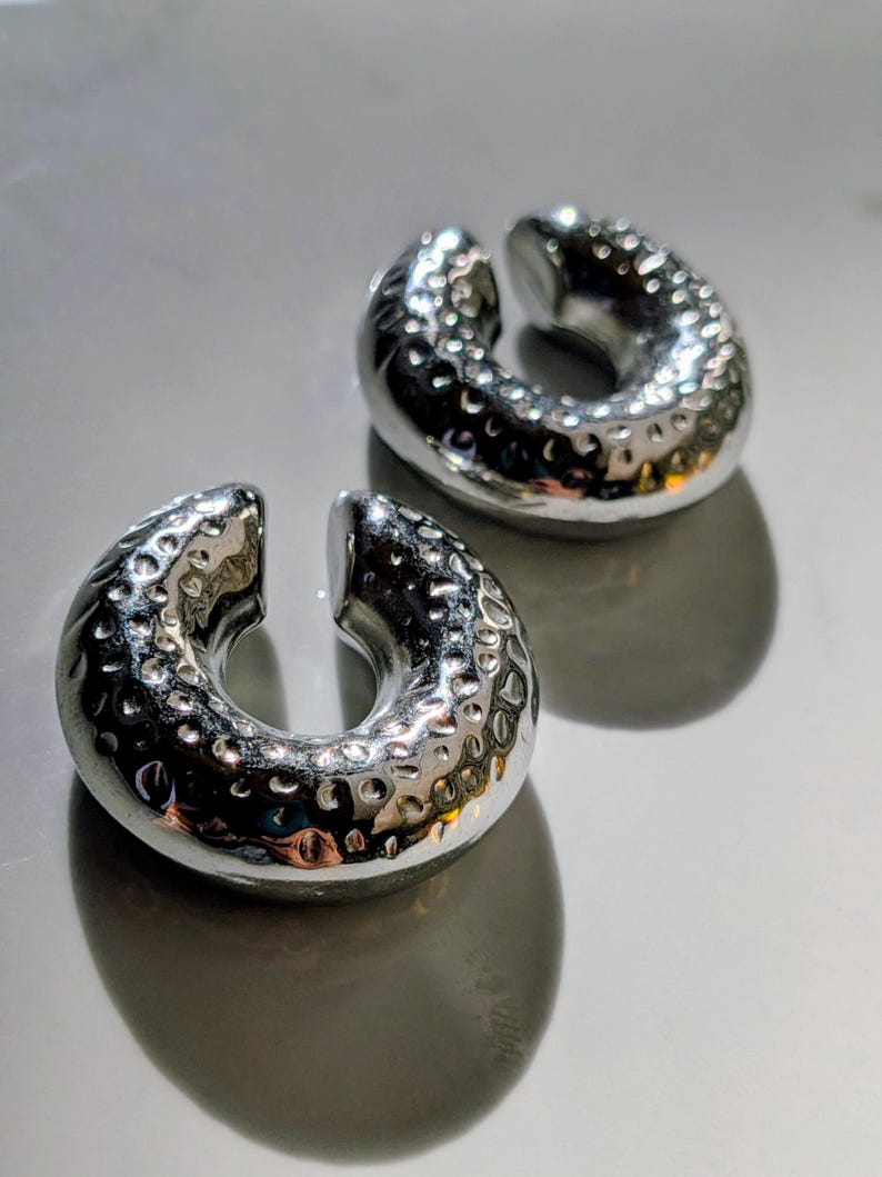 May include: Two silver ear weights with a hammered texture. The weights are circular with a split opening, designed for stretched earlobes. They are displayed on a reflective surface. Each weight is approximately 2.5 cm in diameter.