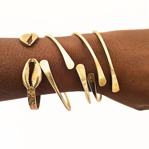 Może przedstawiać: Kolekcja biżuterii w kolorze złotym, w tym pierścionek z motywem muszli, wisiorek w kształcie serca i kilka otwartych bransoletek. Biżuteria jest prezentowana na ramieniu na białym tle.