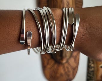 Puede incluir: Un conjunto de pulseras de plata apiladas en la muñeca de una persona. Las pulseras son delgadas y tienen una forma ligeramente curvada.