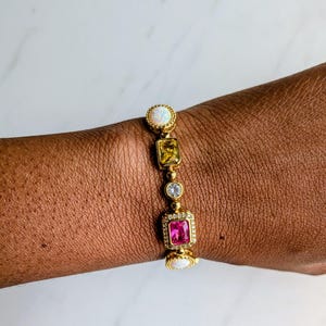 May include: Gold-toned bracelet featuring a series of colorful gemstones. The bracelet includes a white opal, yellow, clear, and pink stones. The bracelet is worn on a wrist, showcasing the jewelry's design and color.