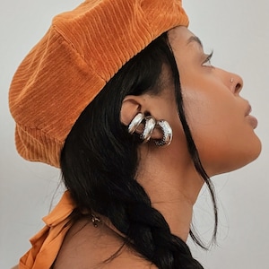 May include: An orange corduroy beret and silver hoop earrings. The beret is a warm, earthy tone, and the earrings have a textured design. The subject has dark hair styled in a braid.