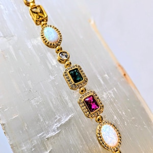 May include: A gold bracelet featuring a series of colorful gemstones in various shapes. The bracelet includes pink, green, yellow, and white stones, with a gold setting and small accent stones. The bracelet is displayed on a white surface.