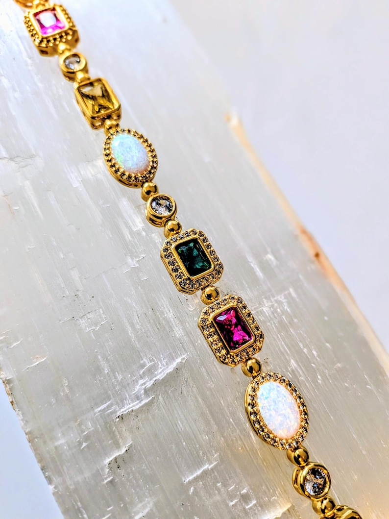 May include: A gold bracelet featuring a series of colorful gemstones in various shapes. The bracelet includes pink, green, yellow, and white stones, with a gold setting and small accent stones. The bracelet is displayed on a white surface.
