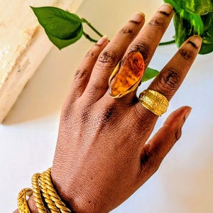 Puede incluir: Una mano adornada con joyas de oro, incluyendo un gran anillo de color ámbar con un engaste de oro, un anillo de banda de oro texturizado y múltiples pulseras de oro. Las joyas se exhiben sobre un fondo neutro.