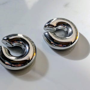 May include: Two polished, silver-coloured ear weights. Each weight has a curved, C-shape design with a smooth, reflective surface. The weights are displayed on a white surface, with shadows adding depth.