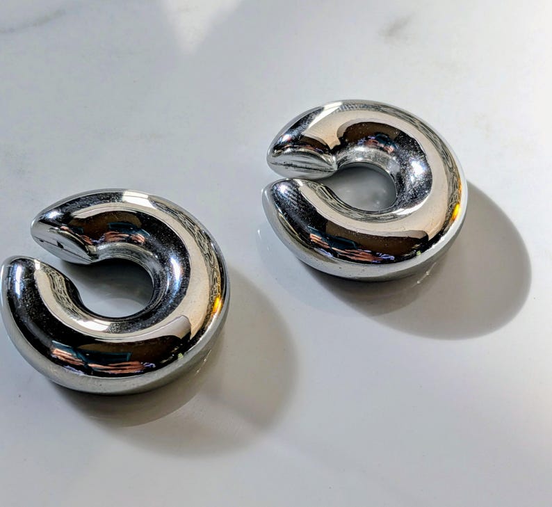 May include: Two polished, silver-coloured ear weights. Each weight has a curved, C-shape design with a smooth, reflective surface. The weights are displayed on a white surface, with shadows adding depth.