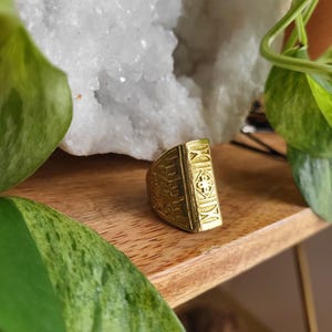 May include: A gold-toned ring with intricate designs and Roman numerals. The ring is sitting on a wooden surface.