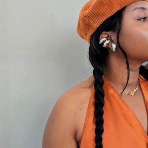 May include: An orange corduroy beret and a matching orange halter top. Silver ear cuffs and a gold necklace with the word "Goddess" are visible. The model has long black hair in a braid.