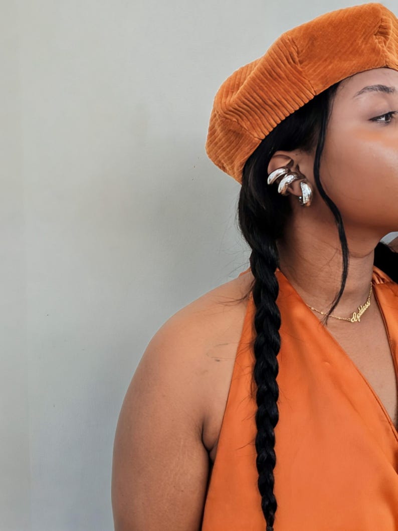May include: An orange corduroy beret and a matching orange halter top. Silver ear cuffs and a gold necklace with the word "Goddess" are visible. The model has long black hair in a braid.