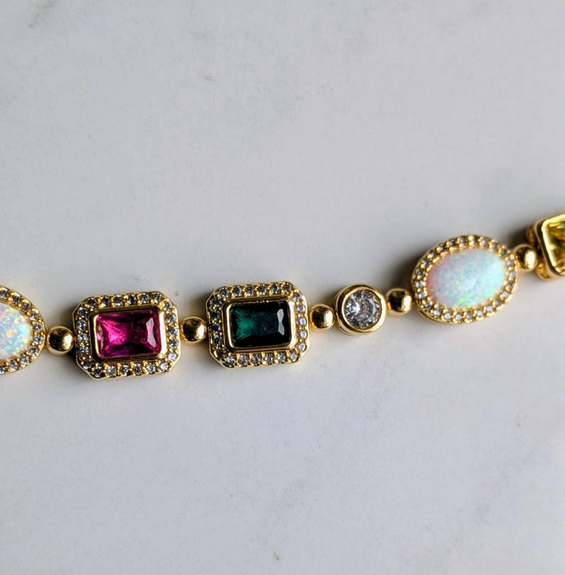 May include: Gold-toned bracelet featuring a series of colorful gemstones. The bracelet includes rectangular, oval, and round stones in shades of pink, green, white, and clear, each set in a gold frame and accented with smaller clear stones.