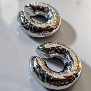 May include: Two shiny, silver ear weights with a textured, scale-like pattern. The weights are crescent-shaped with a split in the middle, designed for stretched ear piercings. The objects are displayed on a light-coloured surface.