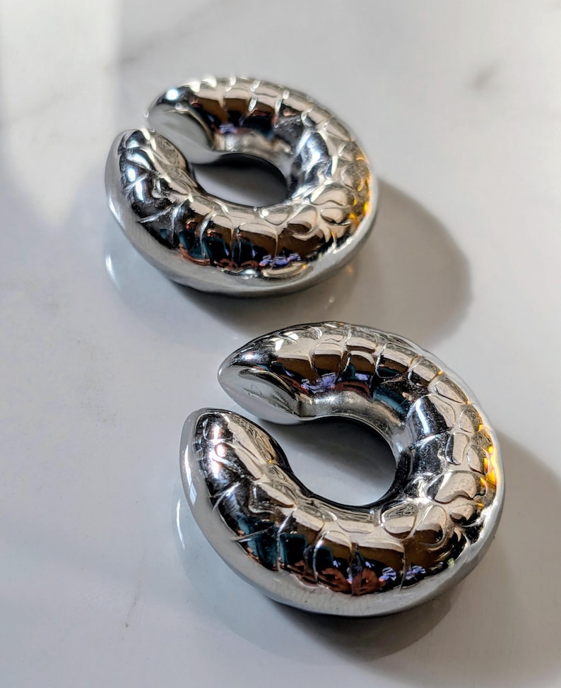 May include: Two shiny, silver ear weights with a textured, scale-like pattern. The weights are crescent-shaped with a split in the middle, designed for stretched ear piercings. The objects are displayed on a light-coloured surface.