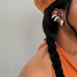 May include: An orange corduroy beret and silver hoop earrings are visible. The earrings have a layered design. The person's hair is styled in a long, dark braid. The top is orange.