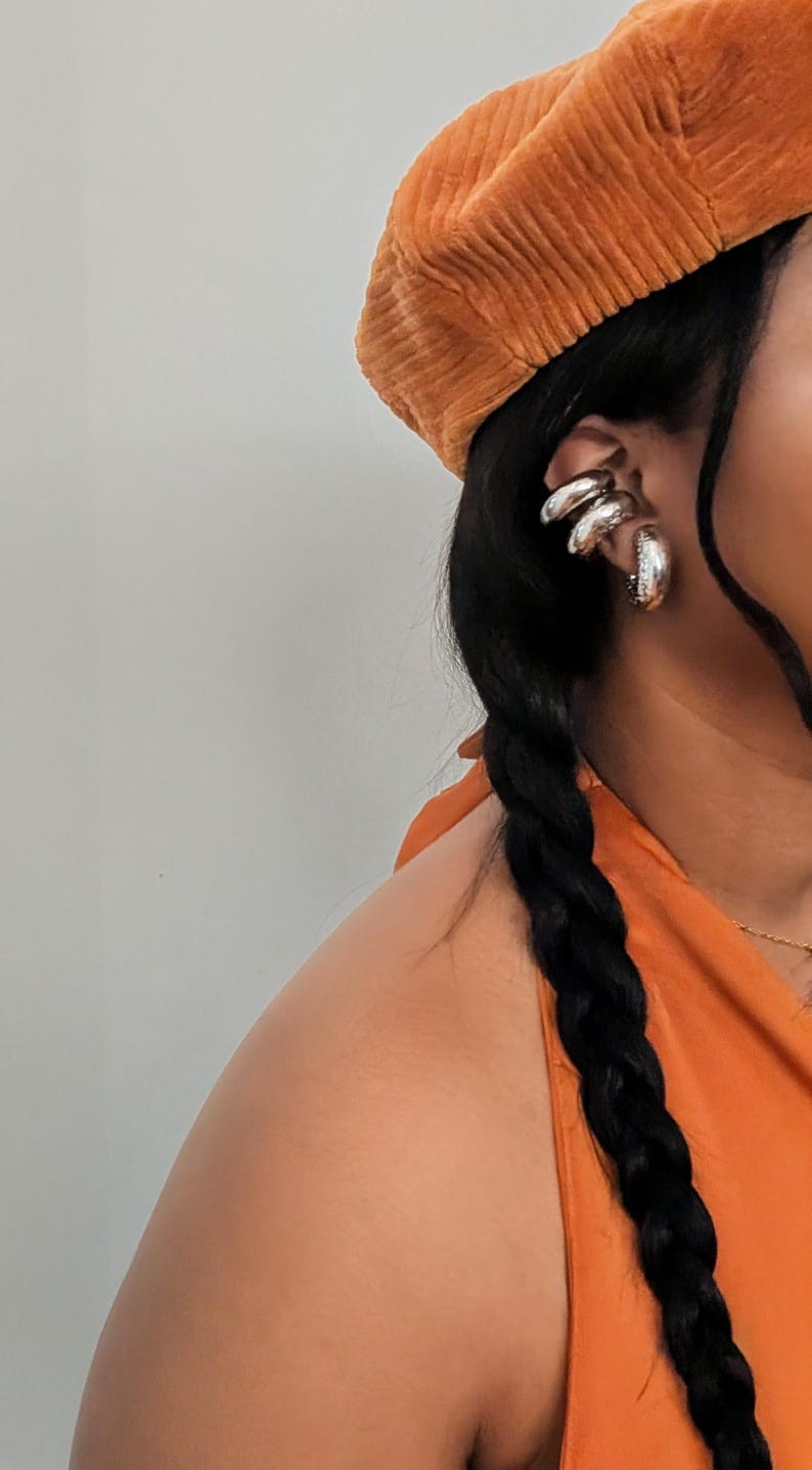 May include: An orange corduroy beret and silver hoop earrings are visible. The earrings have a layered design. The person's hair is styled in a long, dark braid. The top is orange.
