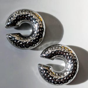 May include: Two silver-coloured, C-shaped ear weights with a hammered texture. The weights have a smooth, polished finish and a circular opening in the centre. The ear weights are displayed on a white surface.
