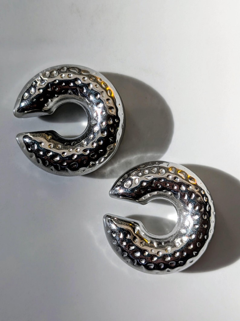 May include: Two silver-coloured, C-shaped ear weights with a hammered texture. The weights have a smooth, polished finish and a circular opening in the centre. The ear weights are displayed on a white surface.
