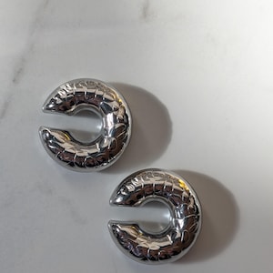 May include: A pair of silver ear cuffs, each shaped like a C with a textured, scale-like surface. The ear cuffs are presented on a white background. These are a contemporary style of ear jewellery.