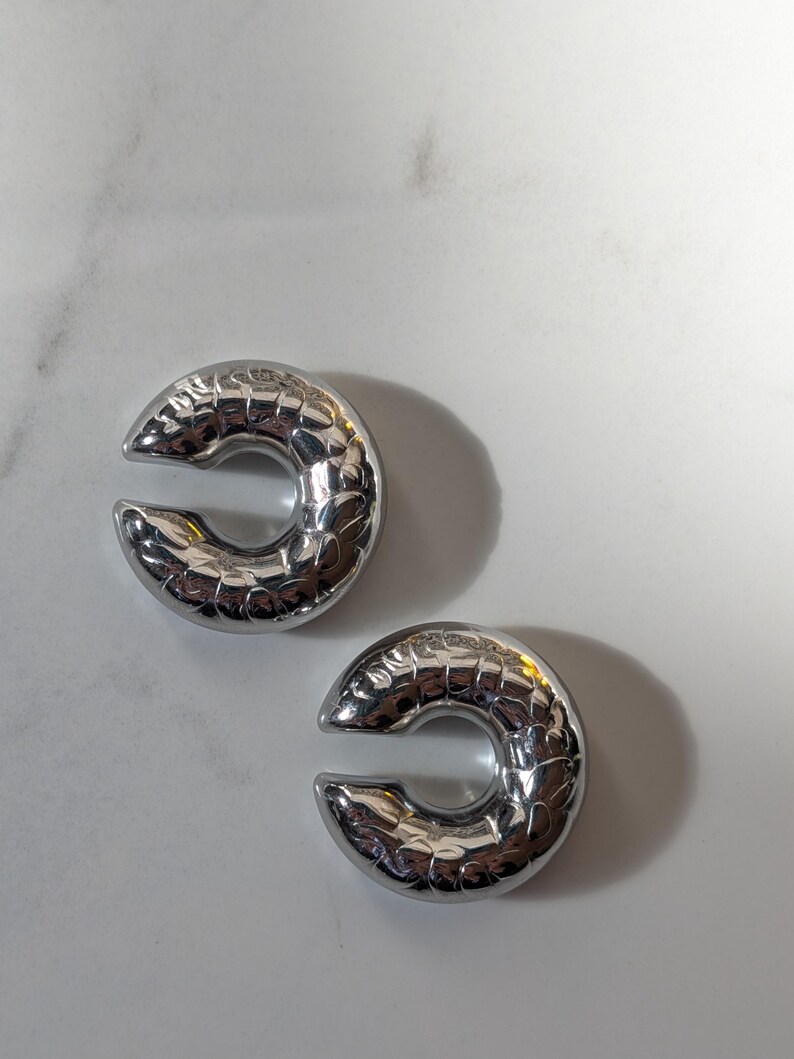 May include: A pair of silver ear cuffs, each shaped like a C with a textured, scale-like surface. The ear cuffs are presented on a white background. These are a contemporary style of ear jewellery.