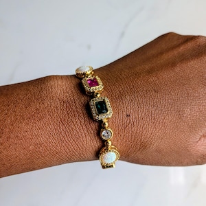 May include: Gold-toned bracelet featuring a series of colorful gemstones. The bracelet includes a white opal, a pink ruby, a green emerald, and a clear diamond-like stone. The bracelet is adorned with small, clear crystals.
