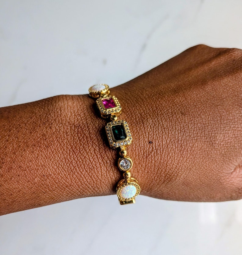 May include: Gold-toned bracelet featuring a series of colorful gemstones. The bracelet includes a white opal, a pink ruby, a green emerald, and a clear diamond-like stone. The bracelet is adorned with small, clear crystals.
