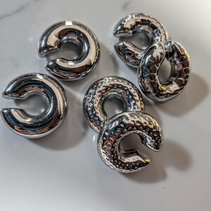 May include: Six shiny, silver-coloured, C-shaped metal objects are displayed. Some have a smooth, reflective surface, while others have a textured, hammered appearance. They are arranged on a white surface, reflecting light.