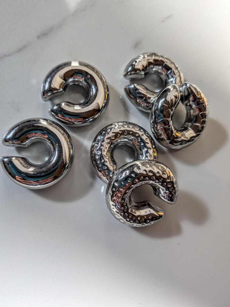 May include: Six shiny, silver-coloured, C-shaped metal objects are displayed. Some have a smooth, reflective surface, while others have a textured, hammered appearance. They are arranged on a white surface, reflecting light.