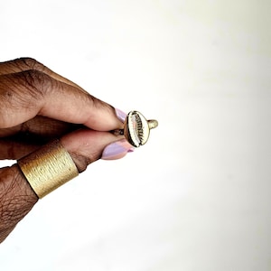 Puede incluir: Un anillo dorado con un diseño de concha de cauri. El anillo tiene una banda texturizada y una pequeña concha detallada. La concha es blanca con acentos marrones y rosas. El anillo se sostiene sobre un fondo blanco.