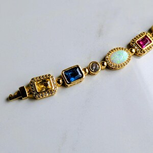 May include: A gold-toned bracelet featuring a variety of colorful gemstones. The bracelet includes a yellow rectangular stone, a blue rectangular stone, a clear round stone, an oval opal, and a red rectangular stone. The stones are set in gold settings.