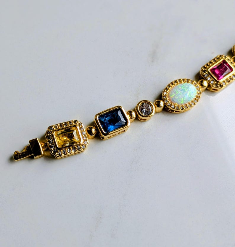 May include: A gold-toned bracelet featuring a variety of colorful gemstones. The bracelet includes a yellow rectangular stone, a blue rectangular stone, a clear round stone, an oval opal, and a red rectangular stone. The stones are set in gold settings.