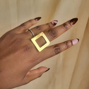 May include: A gold square ring with a hollow center. The ring is worn on the middle finger of a hand with brown nail polish.