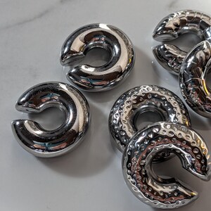 May include: Several shiny, silver-coloured, C-shaped metal objects. Some have a smooth, polished surface, others a textured, hammered finish. The objects are arranged on a light-coloured surface, reflecting the surrounding light.