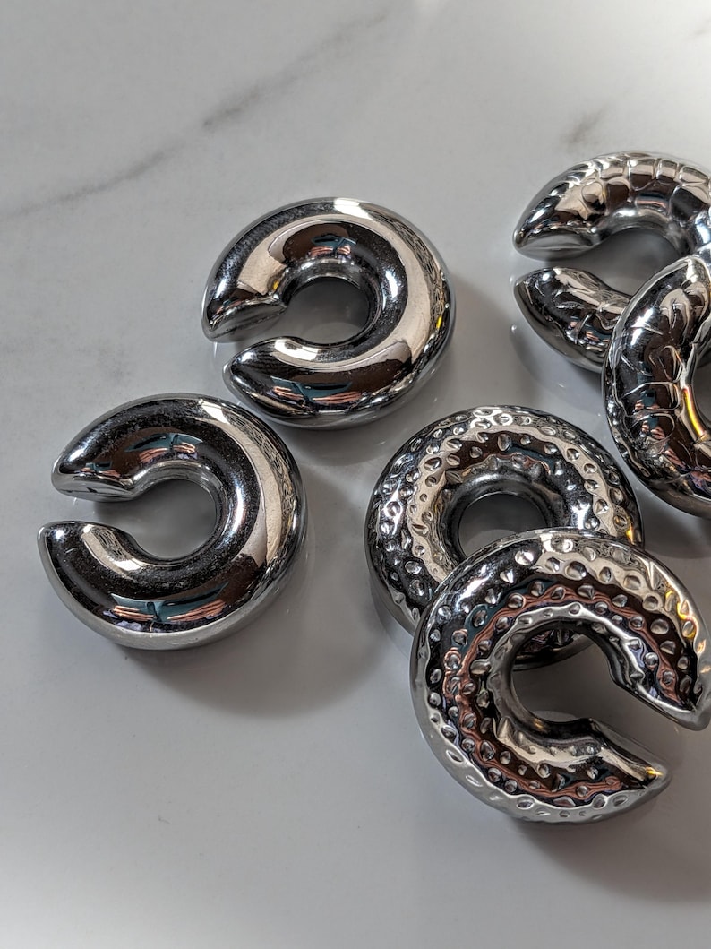 May include: Several shiny, silver-coloured, C-shaped metal objects. Some have a smooth, polished surface, others a textured, hammered finish. The objects are arranged on a light-coloured surface, reflecting the surrounding light.