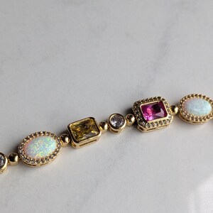 May include: Gold-toned bracelet featuring a variety of gemstones. The bracelet includes oval opals, a yellow square-cut stone, a pink square-cut stone, and clear round stones. The gemstones are set in gold bezels and linked together.