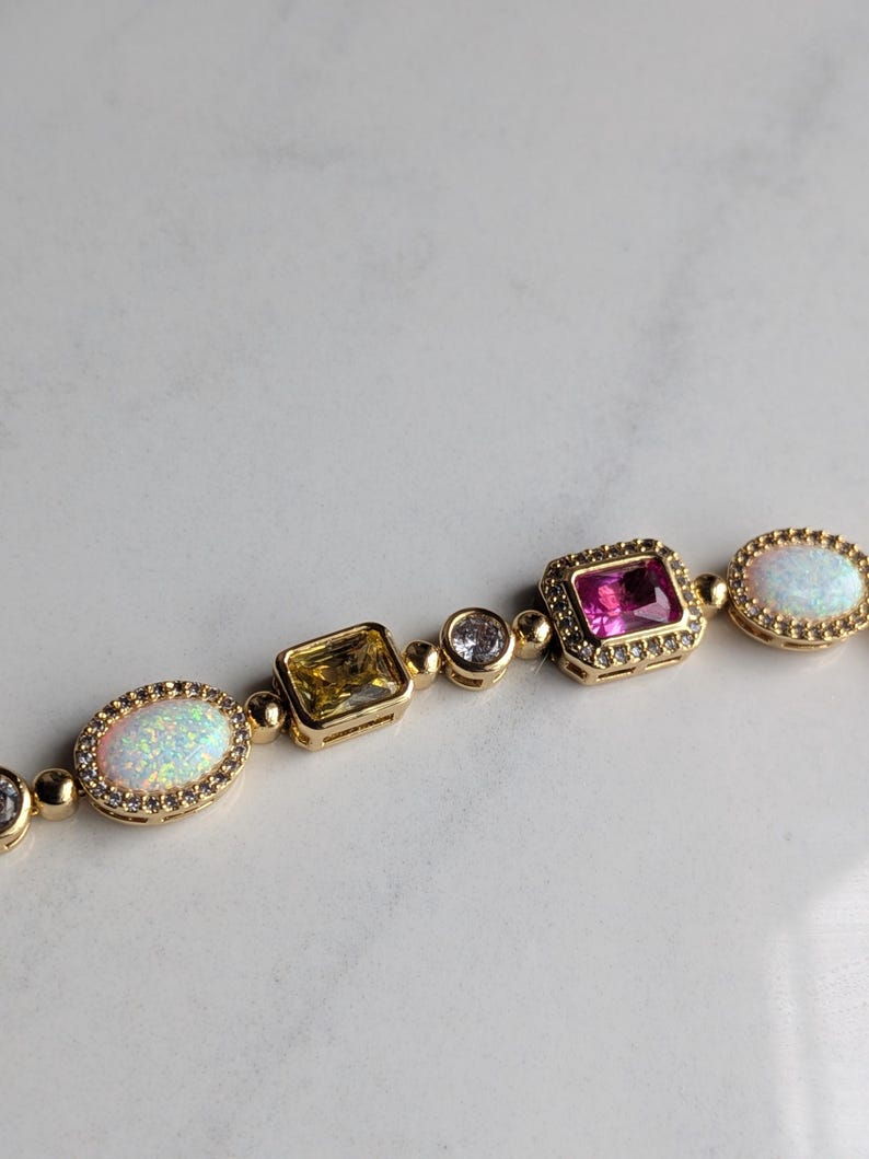 May include: Gold-toned bracelet featuring a variety of gemstones. The bracelet includes oval opals, a yellow square-cut stone, a pink square-cut stone, and clear round stones. The gemstones are set in gold bezels and linked together.