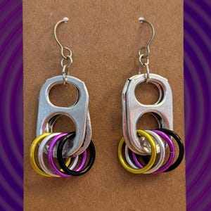 Könnte beinhalten: Ein Paar Ohrringe aus silbernen Pull-Tabs mit gelben, violetten und schwarzen Ringen. Die Ohrringe werden auf einer braunen Karte mit einem Loch oben zum Aufhängen präsentiert.