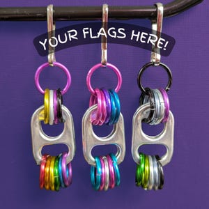 May include: Three keychains made from soda can tabs and colorful rings hang against a purple background. The keychains have pink, black, and silver rings. The text "YOUR FLAGS HERE!" is arched above the keychains.