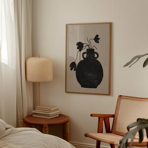 May include: A framed art print featuring a black vase with flowers against a beige background. The vase has two handles and a textured appearance. The artwork is displayed in a room with a lamp, side table, and a chair.