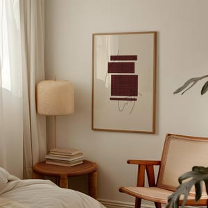 K&ouml;nnte beinhalten: Ein gerahmter abstrakter Druck mit einem burgunderfarbenen geometrischen Design auf cremefarbenem Hintergrund. Der Druck h&auml;ngt an einer wei&szlig;en Wand in einem Raum mit einem Holzstuhl und einer Lampe.