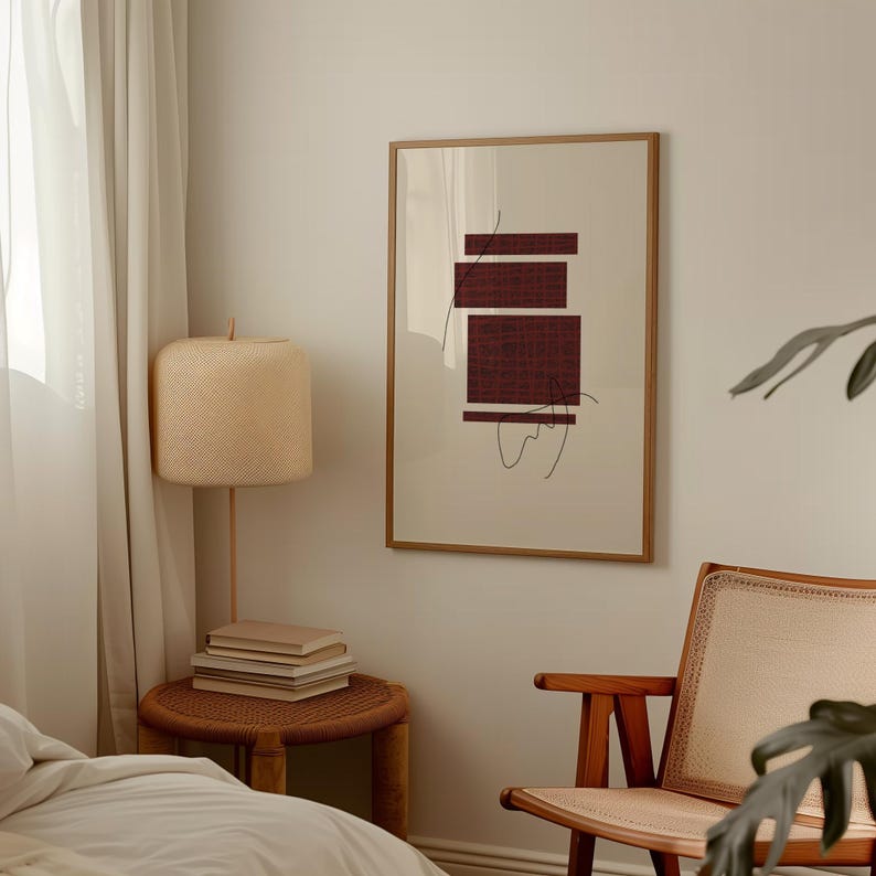 K&ouml;nnte beinhalten: Ein gerahmter abstrakter Druck mit einem burgunderfarbenen geometrischen Design auf cremefarbenem Hintergrund. Der Druck h&auml;ngt an einer wei&szlig;en Wand in einem Raum mit einem Holzstuhl und einer Lampe.