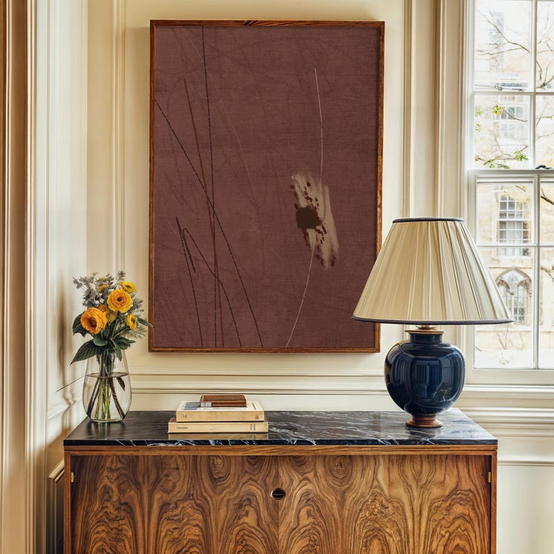 K&ouml;nnte beinhalten: Gerahmtes abstraktes Kunstwerk mit braunem Hintergrund und d&uuml;nnen schwarzen und wei&szlig;en Linien. Eine marineblaue Lampe mit cremefarbenem Schirm steht auf einem Holzschrank mit schwarzer Marmorplatte. Eine Vase mit gelben Blumen steht ebenfalls auf dem Schrank.
