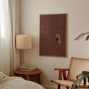K&ouml;nnte beinhalten: Ein gerahmter Kunstdruck mit einem ged&auml;mpften burgunderfarbenen Hintergrund und vertikalen Linien. Eine gewebte Lampe mit beigem Schirm und ein kleiner Beistelltisch mit B&uuml;chern befinden sich im Vordergrund. Rechts ist ein Holzstuhl.