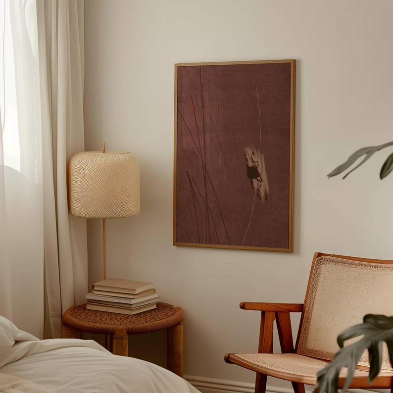 K&ouml;nnte beinhalten: Ein gerahmter Kunstdruck mit einem ged&auml;mpften burgunderfarbenen Hintergrund und vertikalen Linien. Eine gewebte Lampe mit beigem Schirm und ein kleiner Beistelltisch mit B&uuml;chern befinden sich im Vordergrund. Rechts ist ein Holzstuhl.
